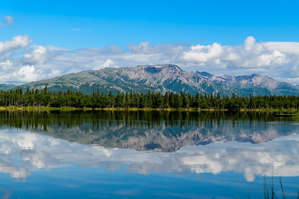 A calm lake reflects a range of rugged, tree-covered mountains beneath a bright blue sky with scattered white clouds, creating a near-mirror image of the landscape along the shoreline.