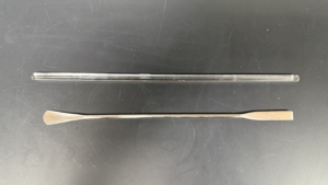 Lab Supplies Glass rod and spatula A clear glass stirring rod and a metal lab spatula with flat ends lying side by side on a black surface. Alternative text created using Copilot and edited by a human on February 26, 2026.