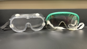 Lab Supplies Safety Goggles Two pairs of safety goggles on a black countertop: one clear pair with a black strap and one green-framed pair with a white strap. Alternative text created using Copilot and edited by a human on February 26, 2026.