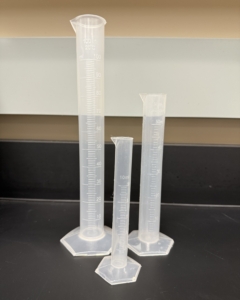 Lab Supplies graduated cylinders Three clear plastic graduated cylinders of different volumes (100mL, 50, mL and 10mL) standing upright on a lab bench, each with measurement markings. Alternative text created using Copilot and edited by a human on February 26, 2026.
