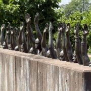 A bronze sculpture displayed outdoors on a sunny day, featuring a row of human figures emerging from the top of a large weathered wooden block. The figures appear to be submerged from the chest down, with only their heads, faces, and raised arms visible above the surface. Each figure's arms are stretched upward with palms open, and some figures wear shackles or chains on their wrists, evoking themes of slavery, captivity, and the struggle for freedom. The faces of the figures convey anguish and resistance. The sculpture is set against a backdrop of lush green trees and a well-maintained grassy lawn, suggesting a memorial or museum setting. This text created using Claude and edited by Catherine Haslag on May 31, 2026.