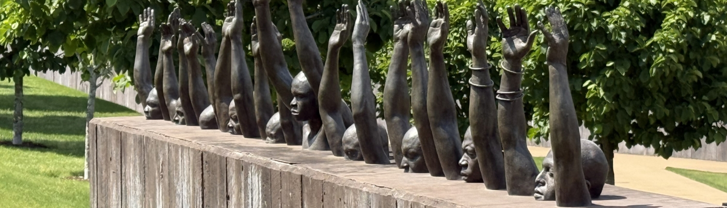 A bronze sculpture displayed outdoors on a sunny day, featuring a row of human figures emerging from the top of a large weathered wooden block. The figures appear to be submerged from the chest down, with only their heads, faces, and raised arms visible above the surface. Each figure's arms are stretched upward with palms open, and some figures wear shackles or chains on their wrists, evoking themes of slavery, captivity, and the struggle for freedom. The faces of the figures convey anguish and resistance. The sculpture is set against a backdrop of lush green trees and a well-maintained grassy lawn, suggesting a memorial or museum setting. This text created using Claude and edited by Catherine Haslag on May 31, 2026.