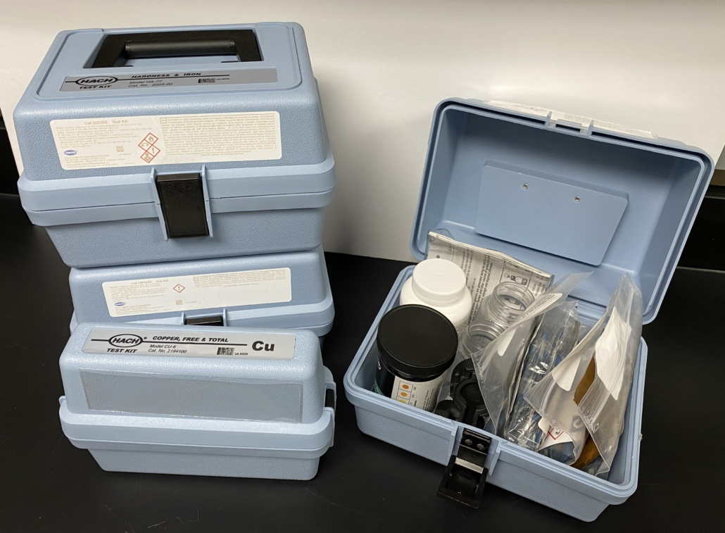 Four light blue HACH kits. Three of the kits are stacked together on the left side of the photo. One is open on the right, displaying bottles and reagent packets.